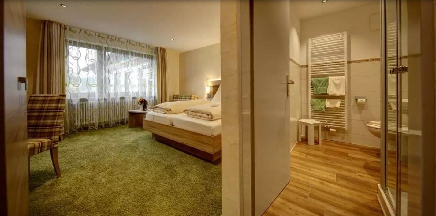 Habitación de hotel con cama, una silla, una mesita con un jarrón y baño con ducha e inodoro.