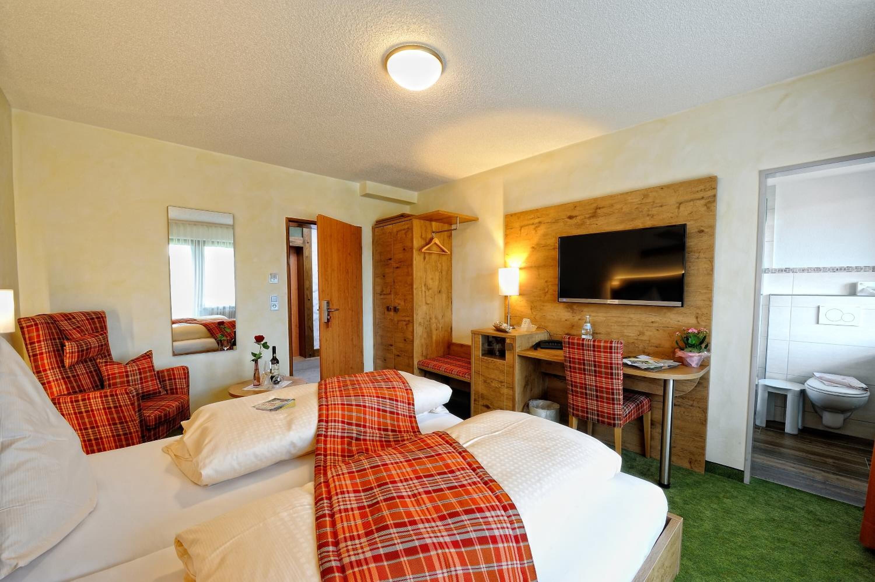A hotel room with a large bed, a desk with a chair, a TV mounted on the wall, and a bathroom visible through an open door.