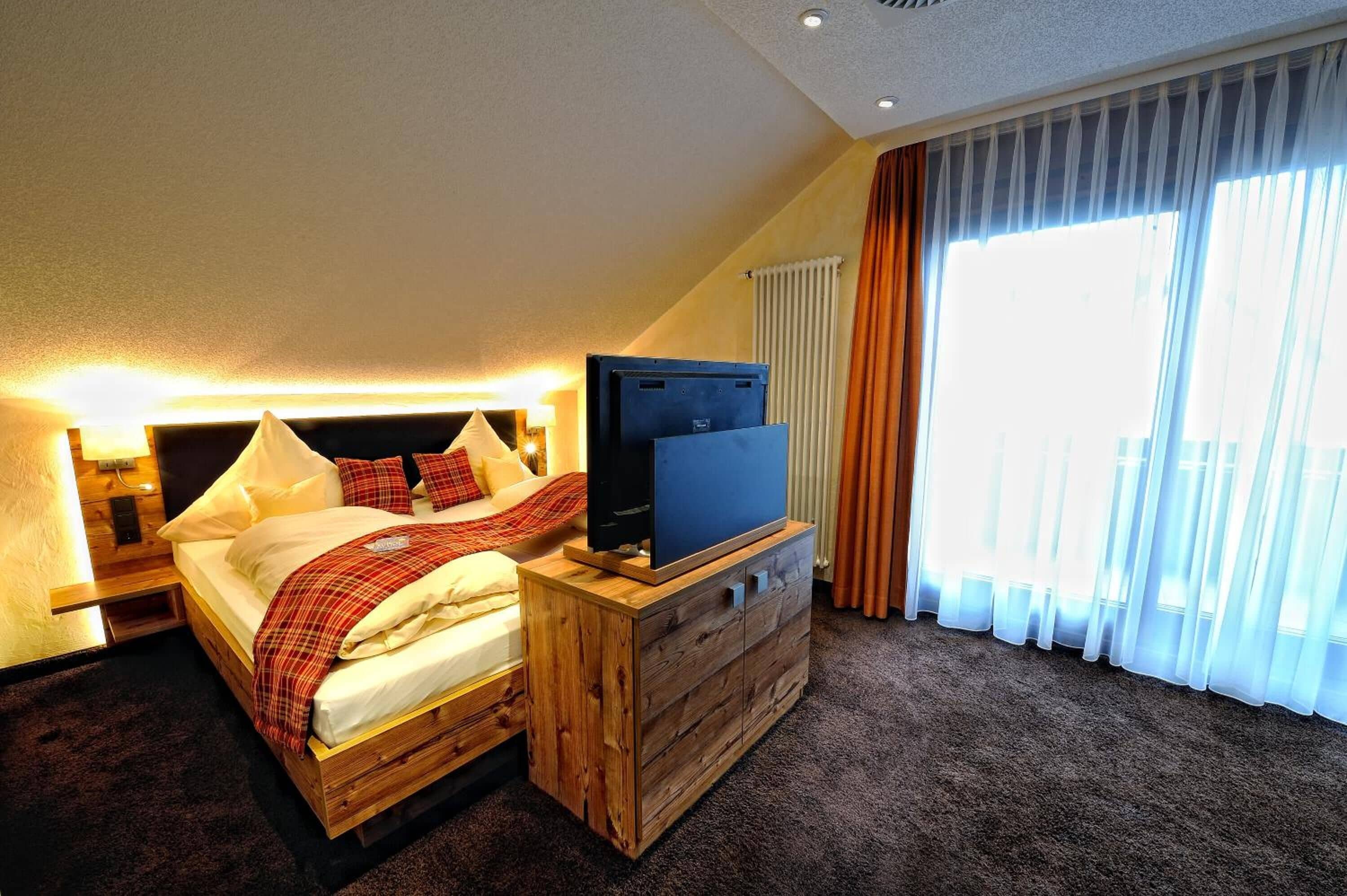 A bedroom with a bed, a TV on a wooden dresser, and a large window with curtains.