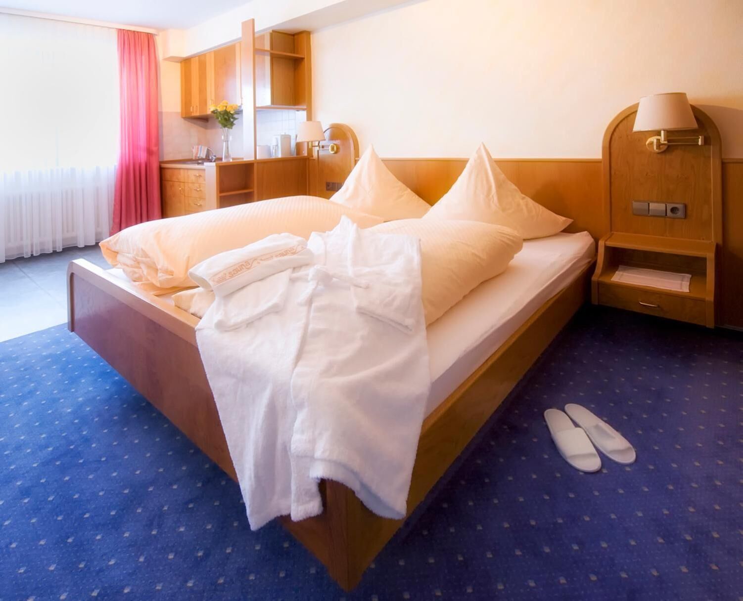 Habitación de hotel con cama, mesita de noche de madera, lámpara y zona de baño con lavabo y espejo.
