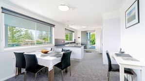 A modern kitchen with a central island, a dining area with a table and chairs, and a view of the outdoors through large windows. - Adrift Apartments (Yamba)