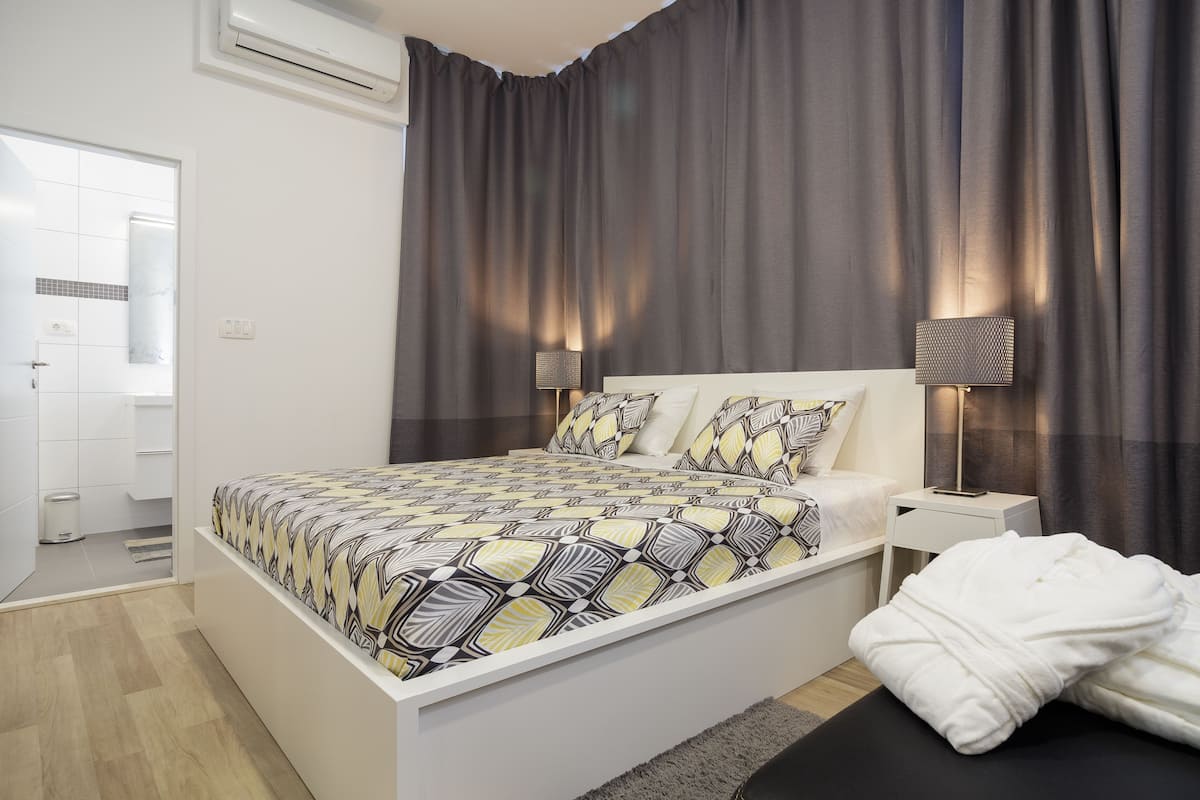 Una habitación de hotel moderna con una cama grande, lámparas de noche, una mesita de noche pequeña y un baño visible a través de una puerta abierta.