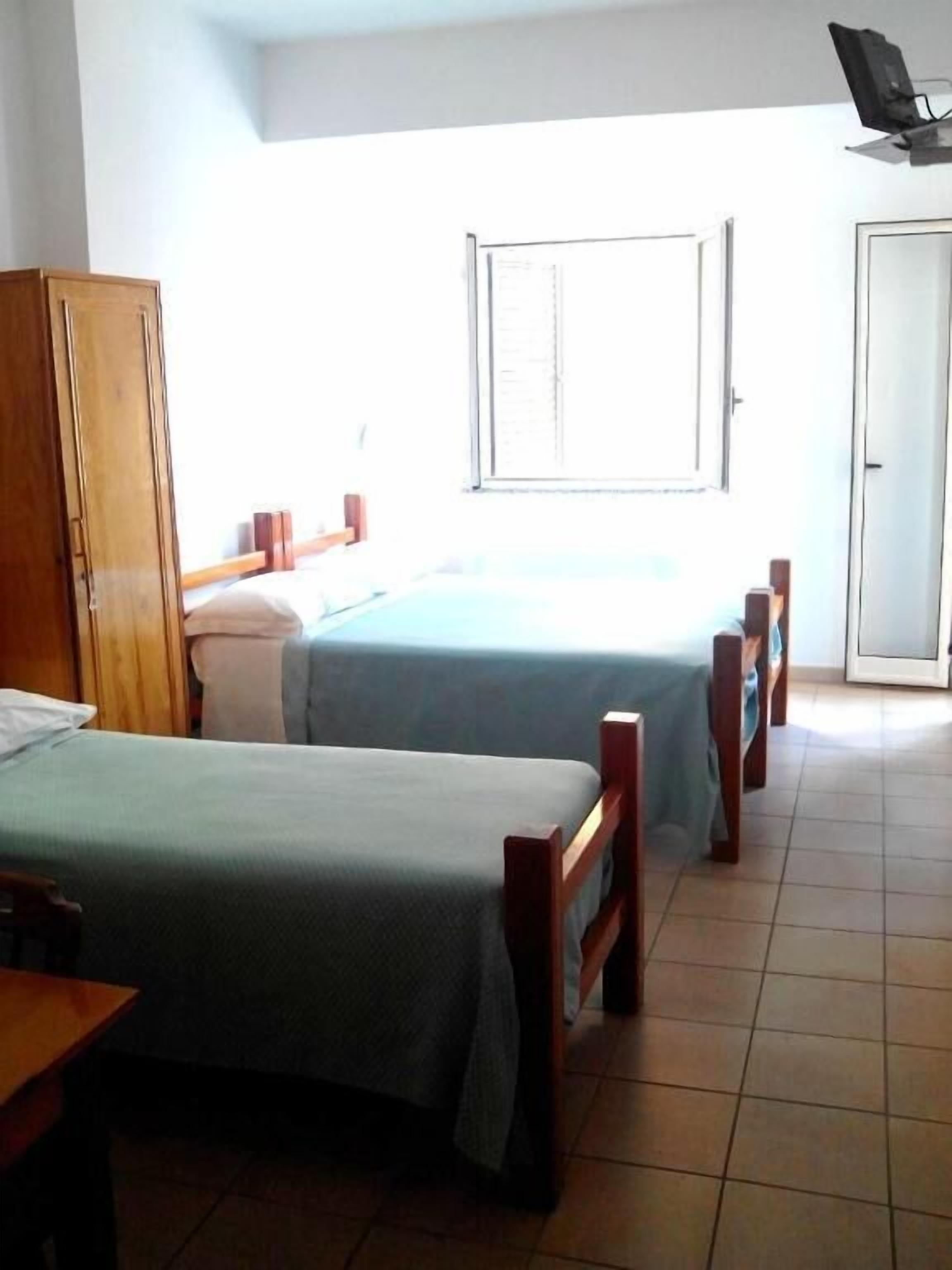 A room with two single beds, a wooden wardrobe, a desk, and a window with blinds.