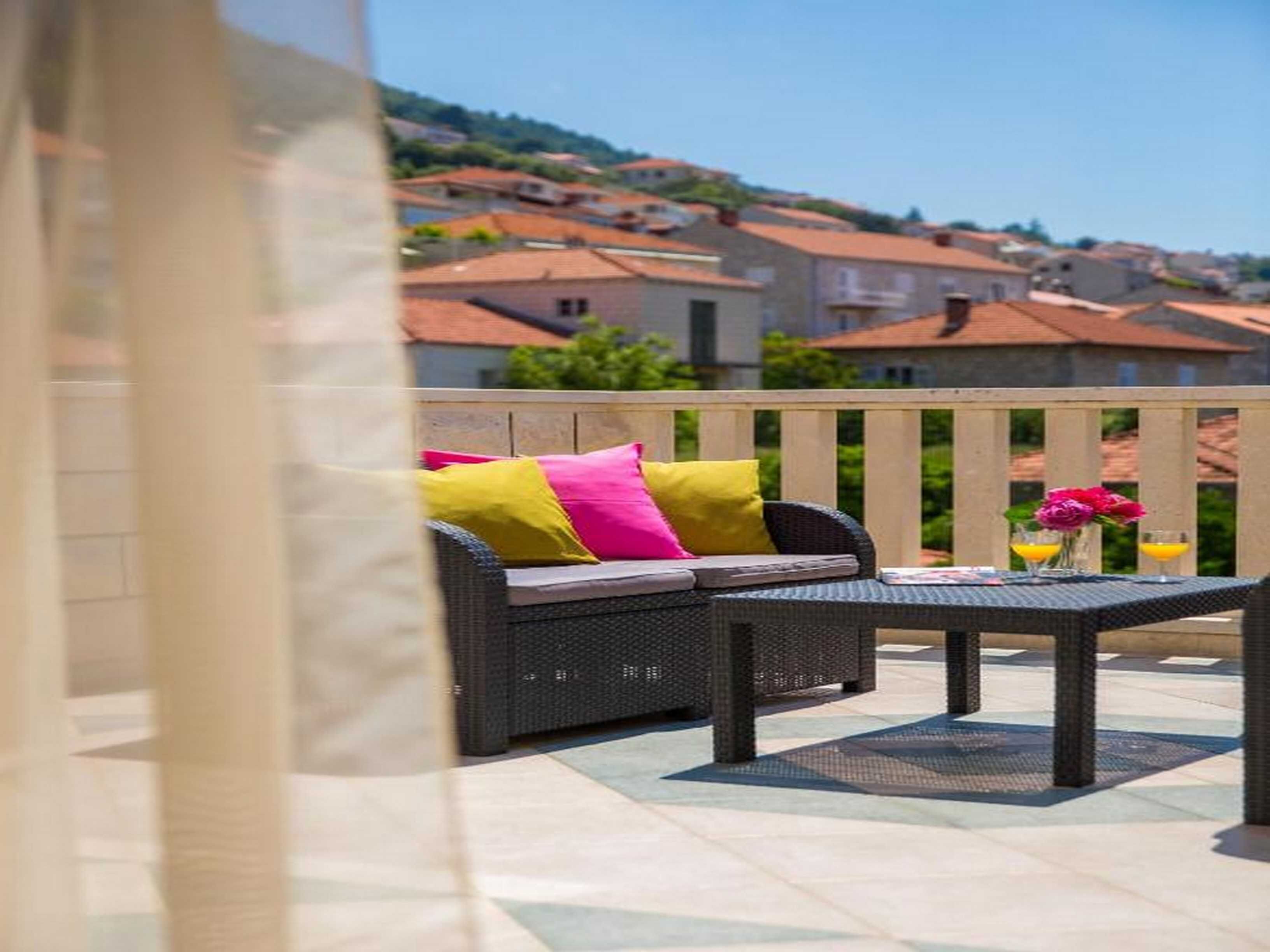 Un patio al aire libre con un sofá de mimbre y una mesa, baranda de madera, y vista a casas al fondo.