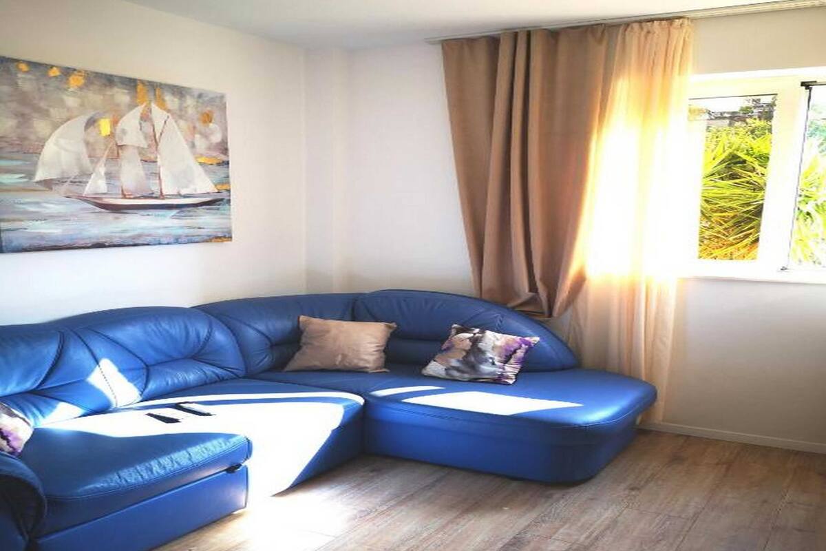 Un salon avec un canapé d’angle bleu, un tableau représentant des voiliers, une fenêtre avec des rideaux et un plancher en bois.