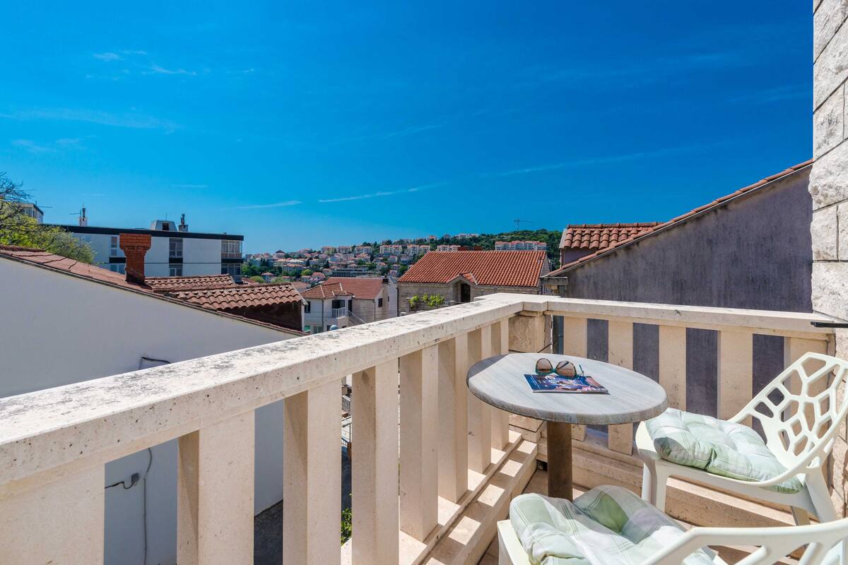 Un balcon avec une table ronde et deux chaises blanches, donnant sur une ville aux toits recouverts de tuiles rouges.