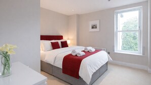 A modern bedroom with a bed, white and red bedding, a bedside table with a vase of flowers, and a window with a view of trees. - Roomspace Apartments -Trinity House (Reigate)