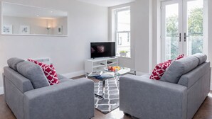 A modern living room with two grey sofas, a glass coffee table, a flat-screen TV, and a large window with a view of greenery. - Roomspace Apartments -Trinity House (Reigate)