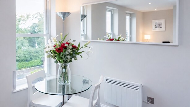 A modern room with a glass table, white chairs, a vase with flowers, a large mirror, and a radiator. - Roomspace Apartments -Trinity House (Reigate)