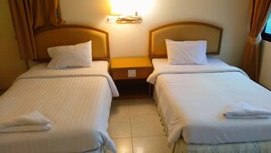 Camera d'albergo con due letti singoli, ognuno con un cuscino e un asciugamano piegato. Un comodino tra i letti ospita una lampada. Le tende sono beige e il pavimento è di colore chiaro.