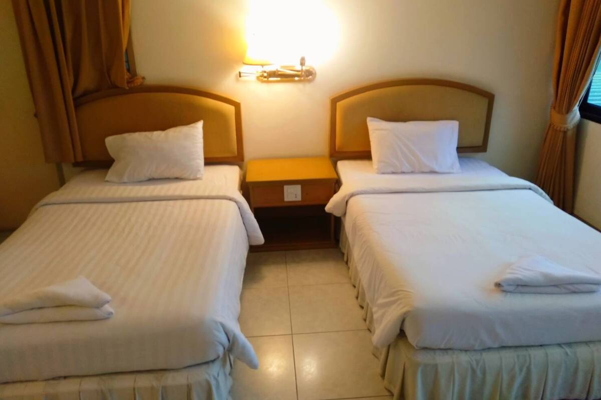 A hotel room with two single beds, each with a pillow and a folded towel. There is a nightstand between the beds with a lamp on it. The room has beige curtains and a light-colored floor.
