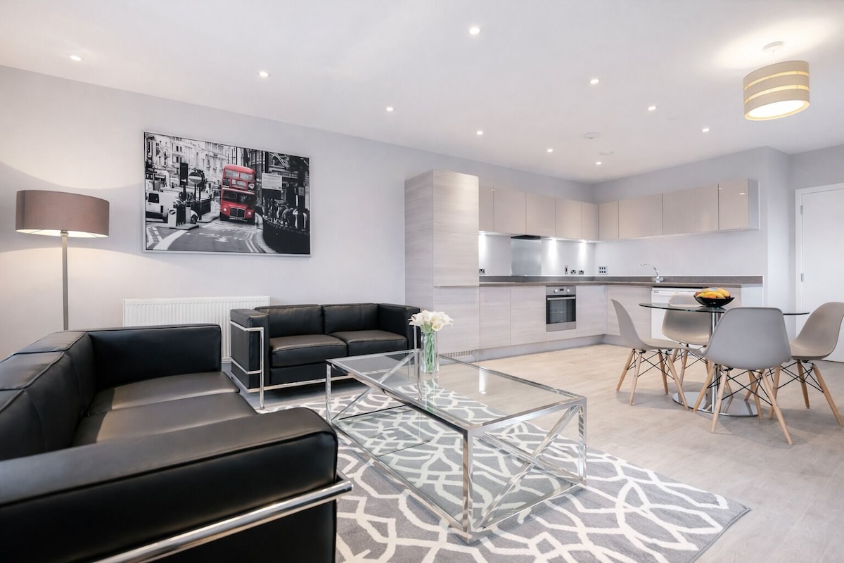 A modern living room with a black leather sofa, a glass coffee table, and a dining area with grey chairs.