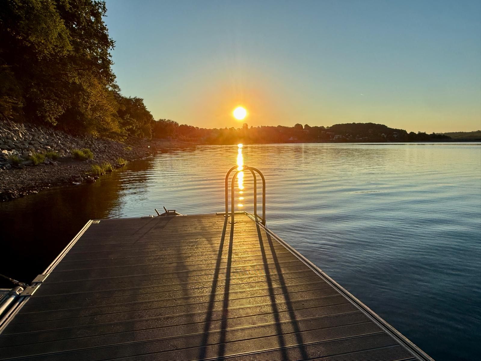 A wooden dock extending into calm water with a metal railing, surrounded by trees and a clear sky.