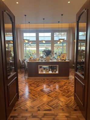 A reception area with a wooden herringbone floor, a reception desk with framed pictures, and large windows with a view of the outdoors. - Haus Delecke (Moehnesee)