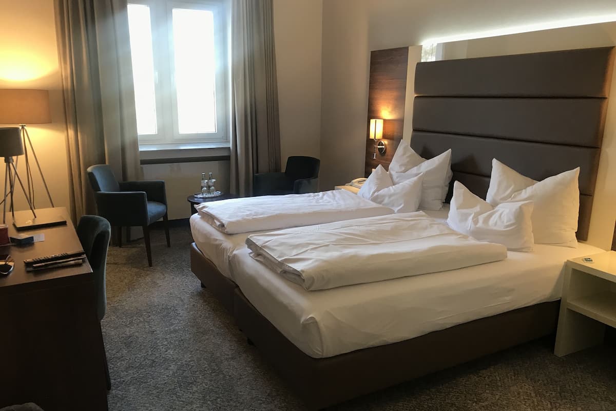 A hotel room with two beds, a desk, a chair, a lamp, and a window with curtains.