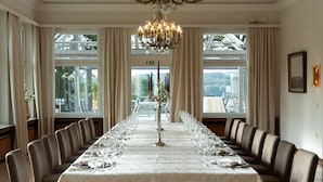 Una larga mesa de comedor dispuesta para una comida formal, rodeada de sillas, con un candelabro colgante y grandes ventanales que permiten la entrada de luz natural.