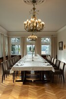 Mesa de jantar longa posta para uma refeição formal, rodeada por cadeiras, com um candelabro acima e grandes janelas permitindo entrada de luz natural.