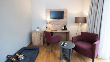 A compact hotel room with a wooden desk, two chairs, a TV mounted on the wall, a small table, a sofa, a lamp, and a pet bed.
