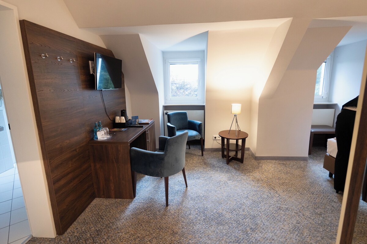 A modern hotel room with a wooden wardrobe, a desk with a chair, a small round table with a lamp, and two armchairs.