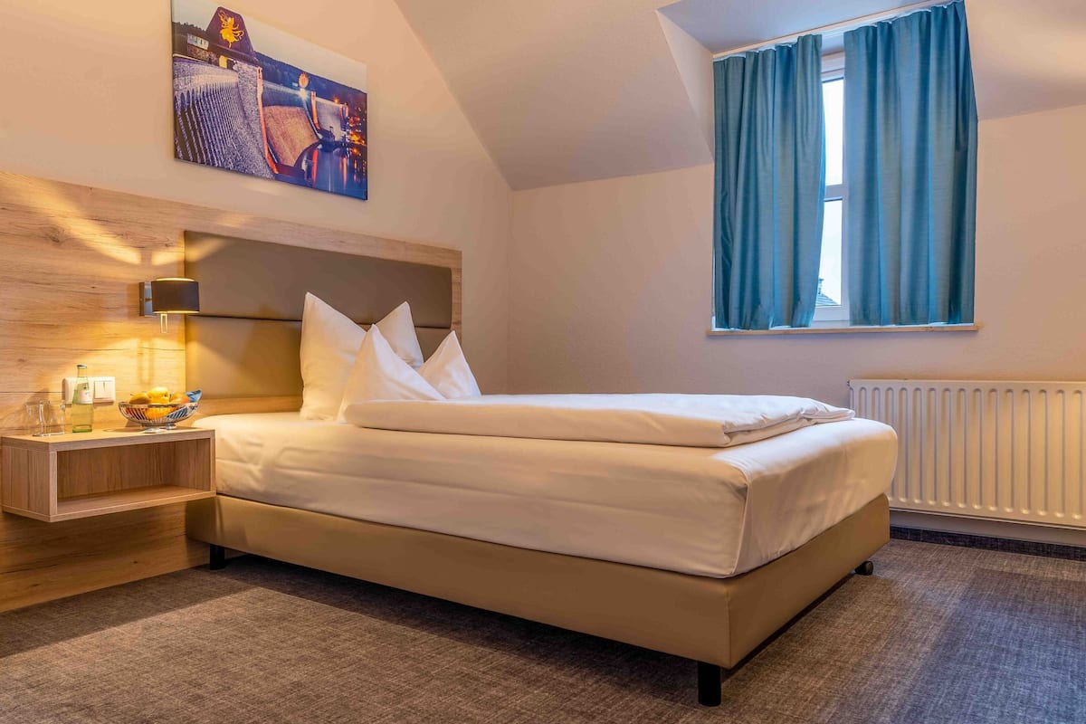 A hotel room with a bed, a nightstand, a lamp, a window with curtains, and a painting on the wall.