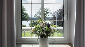 Una ventana con vista a un lago y follaje verde, un jarrón blanco con flores y una cortina blanca.
