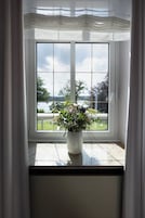 Janela com vista para um lago e vegetação, um vaso branco com flores e uma cortina branca.