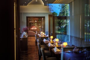 An indoor dining area with tables set for service, a bar area with bottles, and a view of trees through large windows.