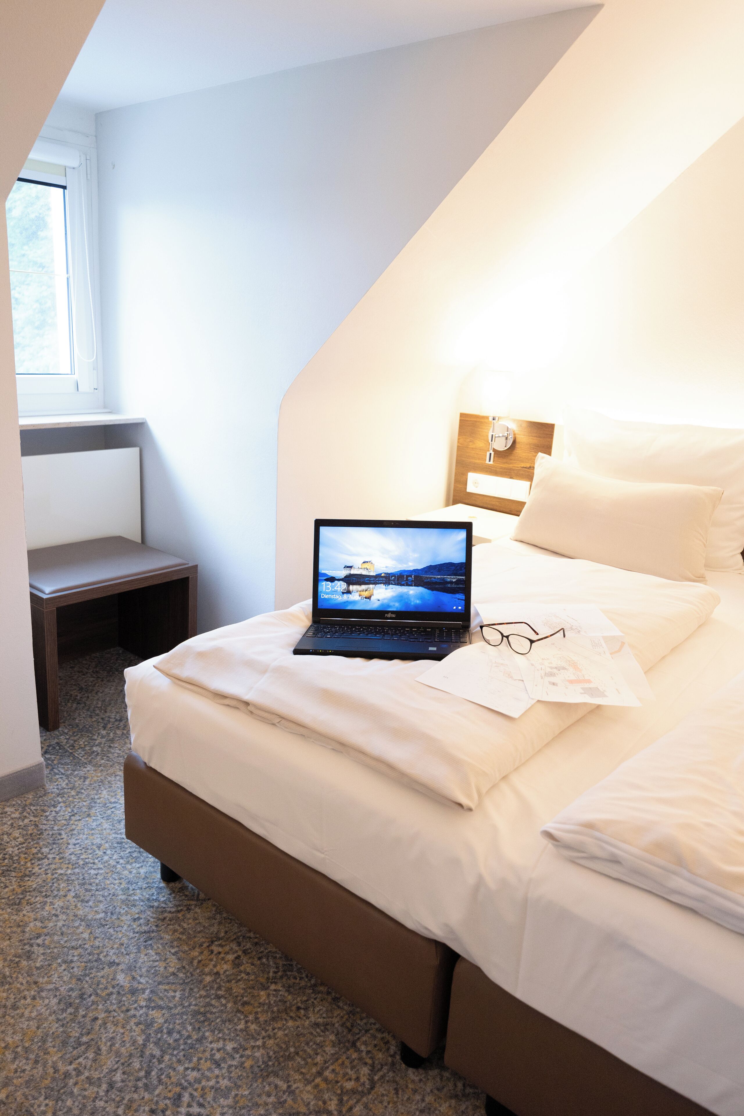 Ein ordentlich bezogenes Bett mit Laptop und Brille darauf, ein kleiner Tisch und ein Fenster in einem Raum mit geneigter Decke.