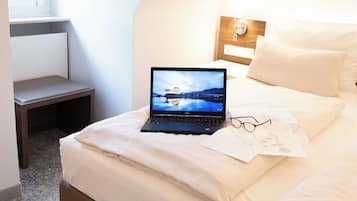 Ein ordentlich bezogenes Bett mit Laptop und Brille darauf, ein kleiner Tisch und ein Fenster in einem Raum mit geneigter Decke.