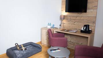 A modern living room with a wooden TV wall, a grey pet bed, two red armchairs, a small round table, and a wooden cabinet with a flat-screen TV.