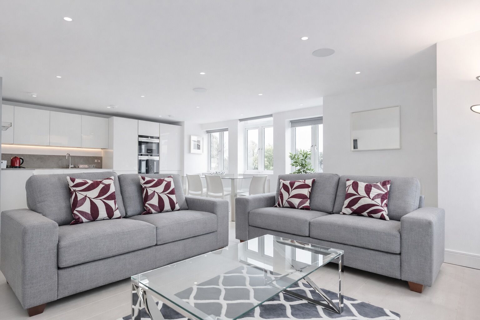 A modern living room with two grey sofas, a glass coffee table, and a kitchen area with white cabinets and a red kettle.