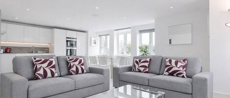 A modern living room with two grey sofas, a glass coffee table, and a kitchen area with white cabinets and a red kettle.
