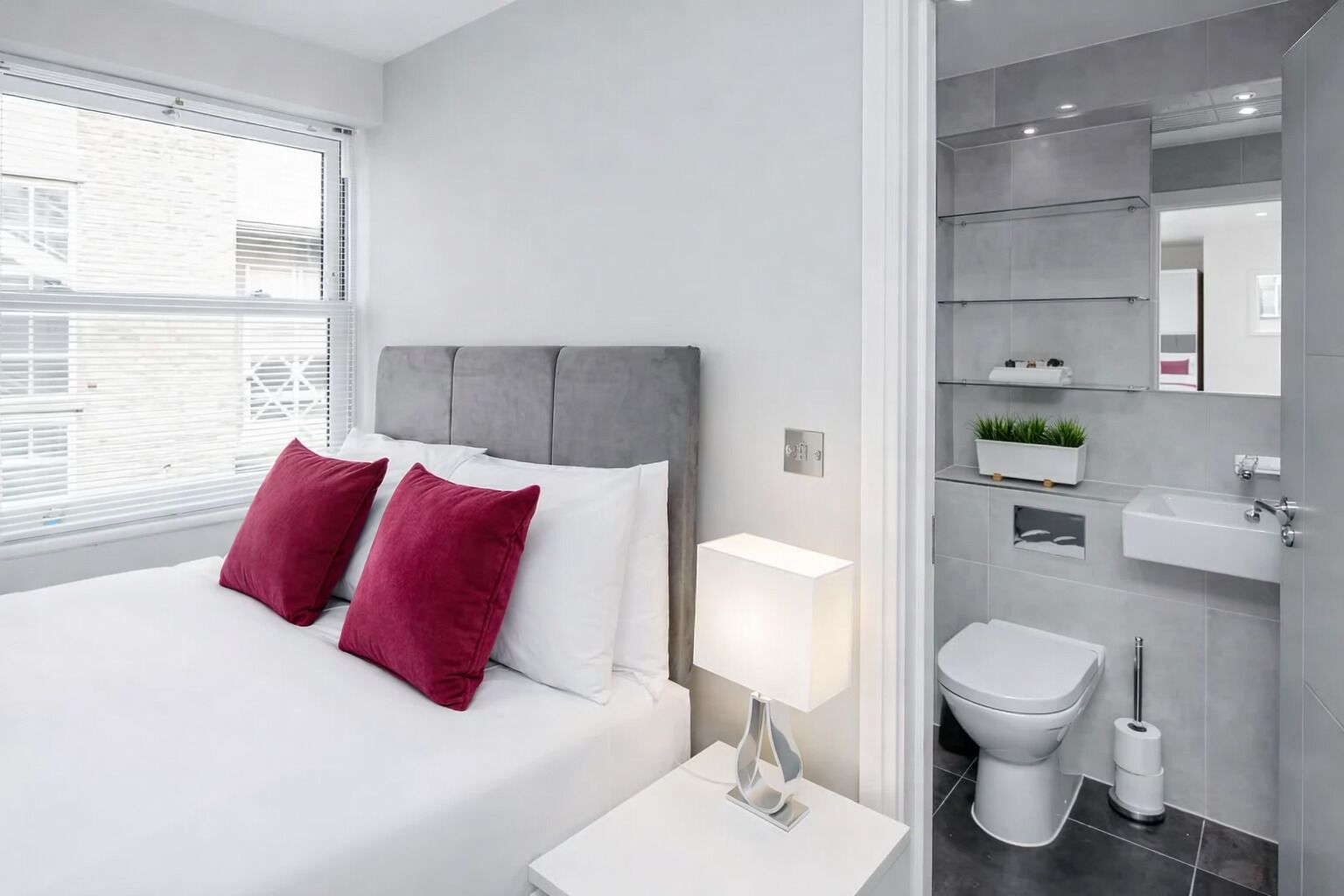 Une chambre d’hôtel moderne avec un grand lit, une table de chevet avec une lampe, et une salle de bain équipée de toilettes, d’un lavabo et d’un miroir.