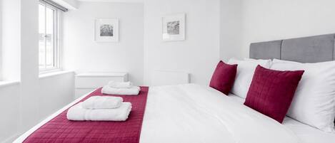 Une chambre d’hôtel moderne avec un grand lit, du linge de lit blanc, une courtepointe rouge matelassée et deux oreillers. Deux tableaux encadrés ornent le mur, il y a une fenêtre avec des stores et une table de chevet blanche.