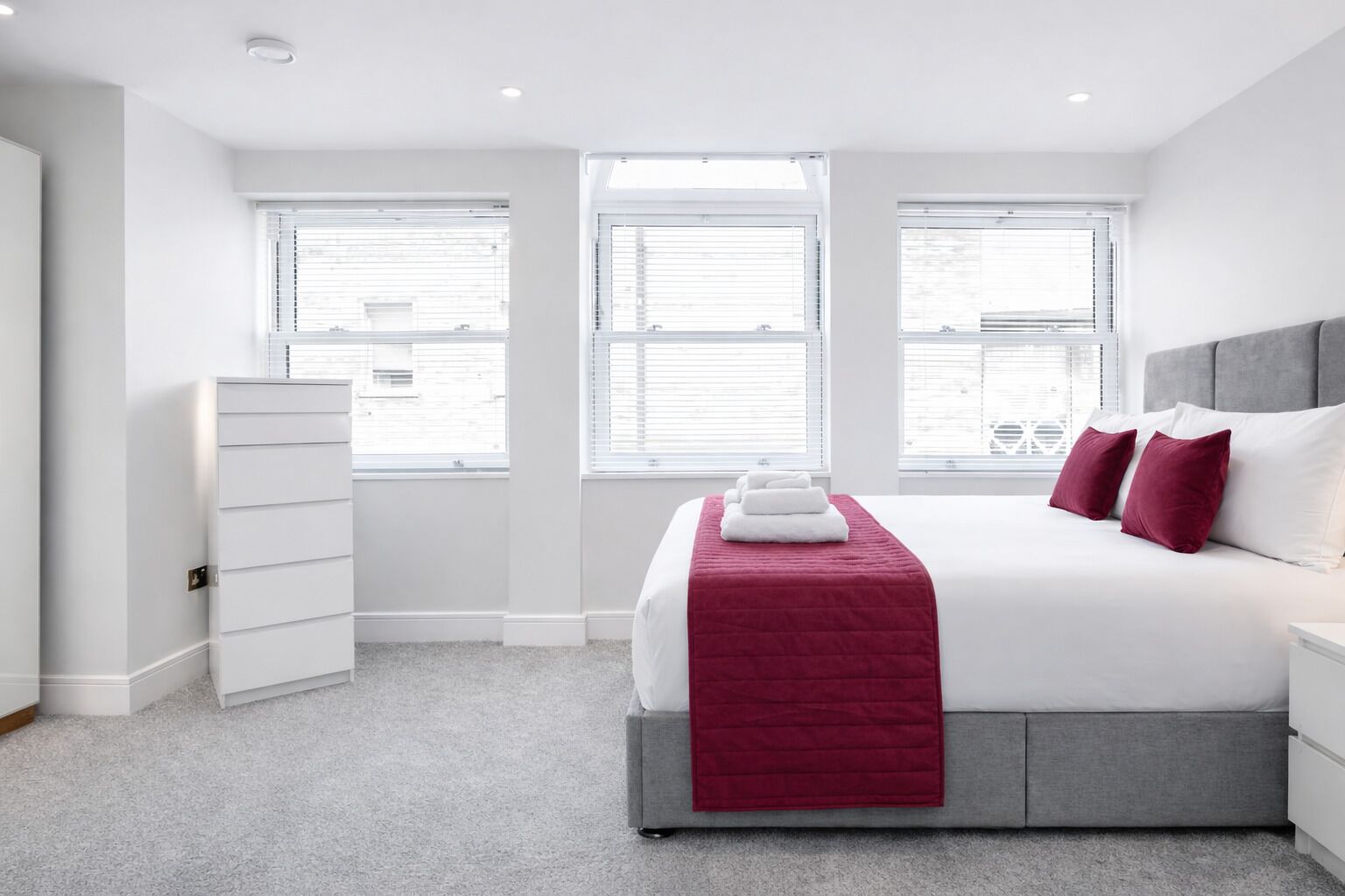 Een moderne hotelkamer met een groot bed, een grijs hoofdeinde en een rode plaid. Twee ramen met jaloezieën, een witte dressoir en een grijs tapijt.