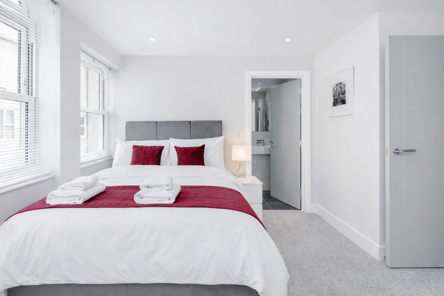 A modern hotel room with a large bed, white bedding, red pillows, and a bedside table with a lamp.