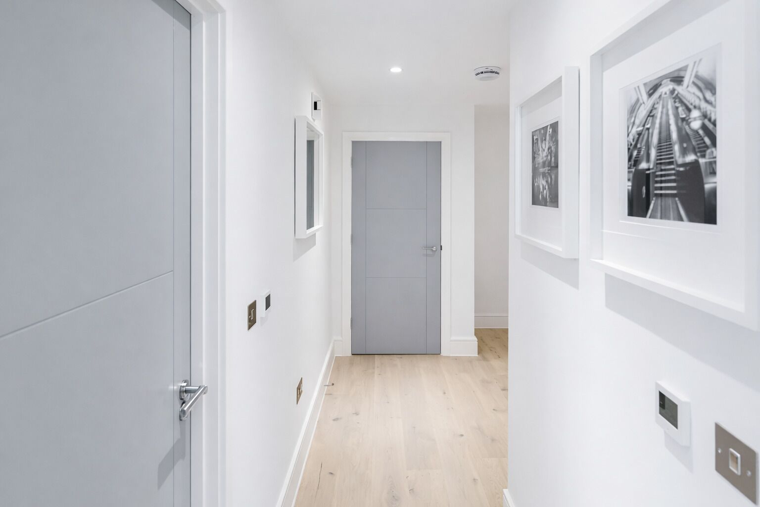 A hallway with white walls, a grey door, and a light wooden floor. There are framed pictures on the walls and a light switch on the wall.