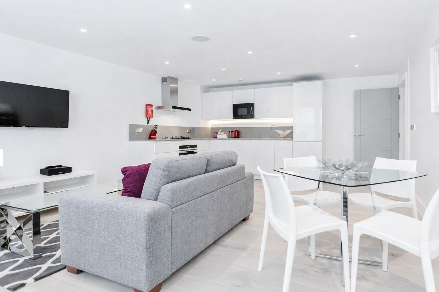 A modern living room with a grey sofa, a glass dining table with white chairs, a flat-screen TV, and a kitchen area with white cabinets and a microwave.