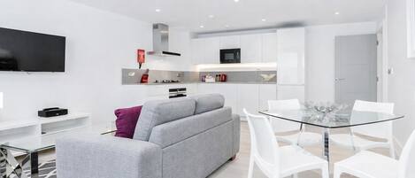 A modern living room with a grey sofa, a glass dining table with white chairs, a flat-screen TV, and a kitchen area with white cabinets and a microwave.