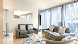 A modern living room with two sofas, a glass coffee table, and a large window with curtains. - Roomspace Apartments -Watling Street (London)