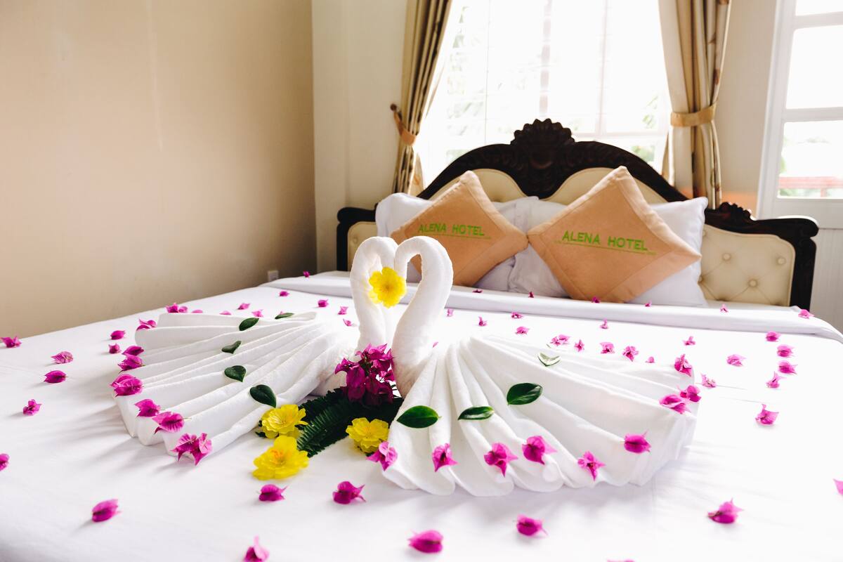 A bed with a swan towel arrangement, pillows with "ALENA HOTEL" branding, and scattered flower petals.
