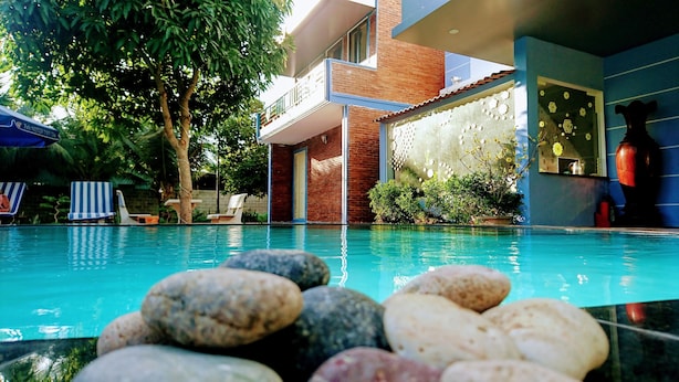 A swimming pool with rocks in the foreground, a brick building with a balcony, and lounge chairs under a tree. - Mui Ne Alena Bungalow Hotel (Mui Ne)