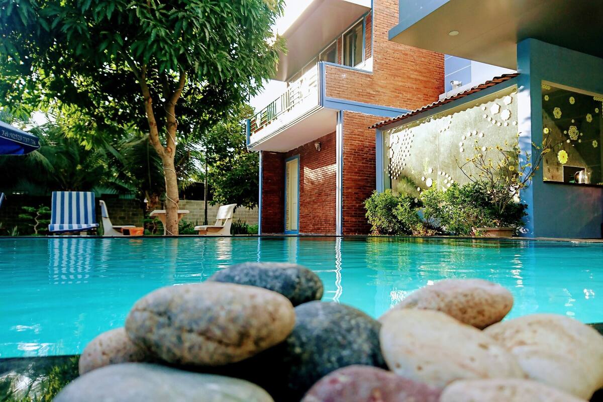 A swimming pool with rocks in the foreground, a brick building with a balcony, and lounge chairs under a tree.