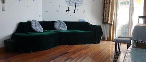 A room with a wooden floor, a green sofa, a chandelier, and a window with curtains.