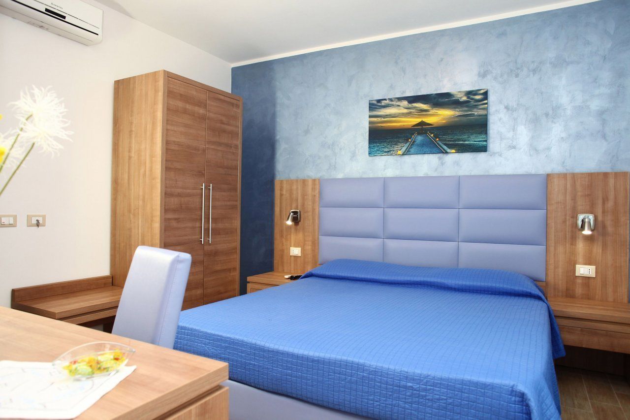 A bedroom with a bed, a wooden wardrobe, a desk with a chair, and a wall-mounted painting of a sunset over the ocean.