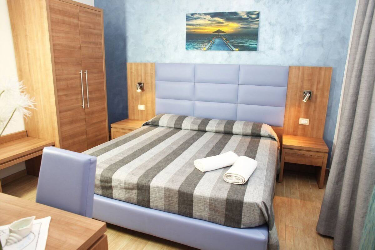 Une chambre d’hôtel avec un lit, une armoire en bois, une table de chevet, un bureau et un tableau représentant un coucher de soleil sur l’océan.