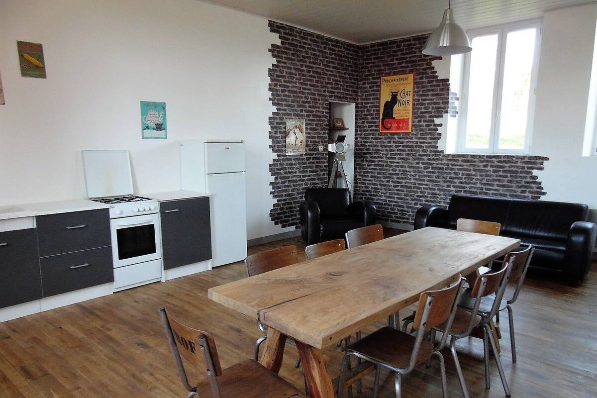 A compact kitchen with a stove, refrigerator, and white cabinets. A dining table with chairs is in the center, and a black leather sofa is against the brick wall.