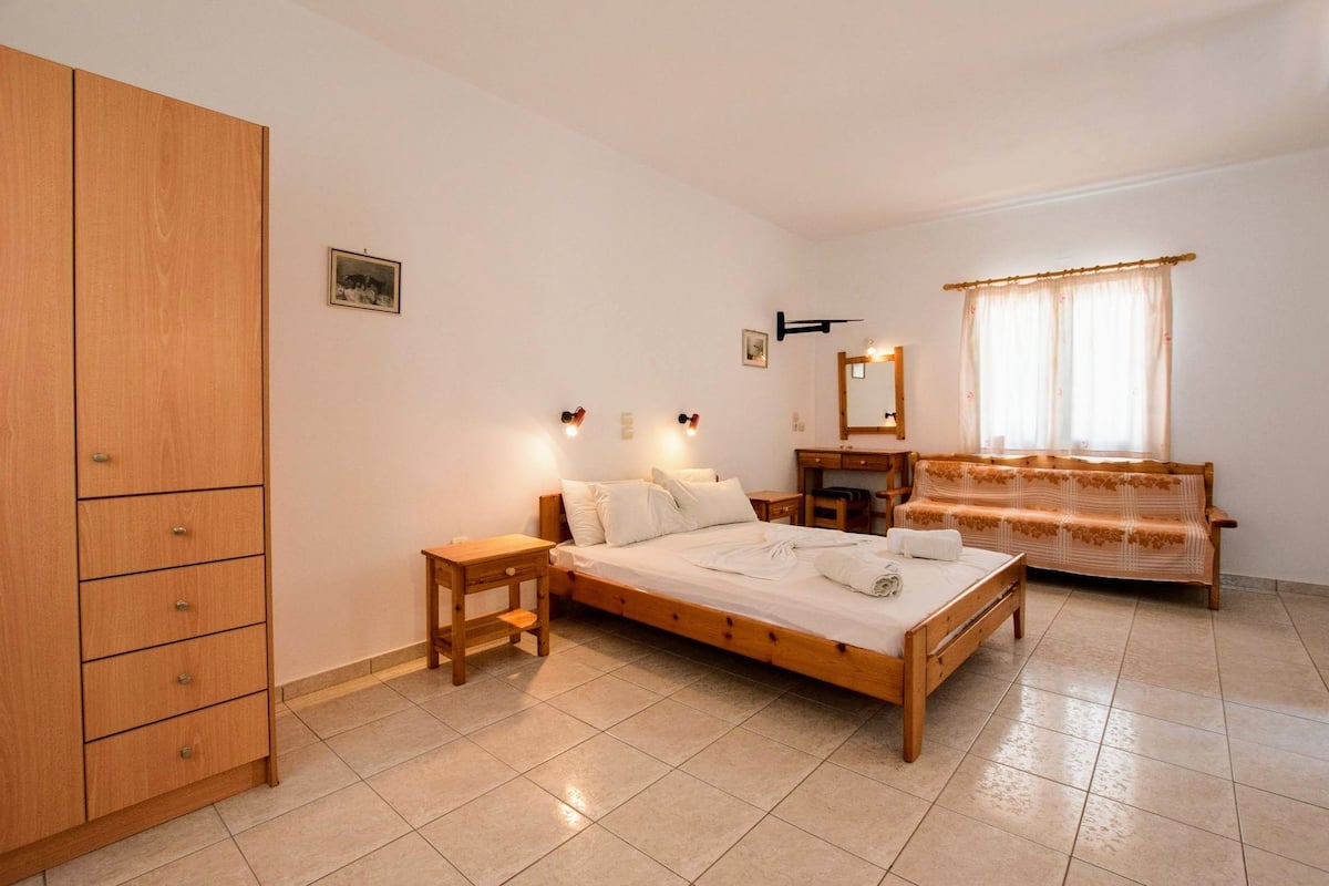 A bedroom with a wooden bed, a wooden wardrobe, a bedside table, a bench, a mirror, and a window with curtains.