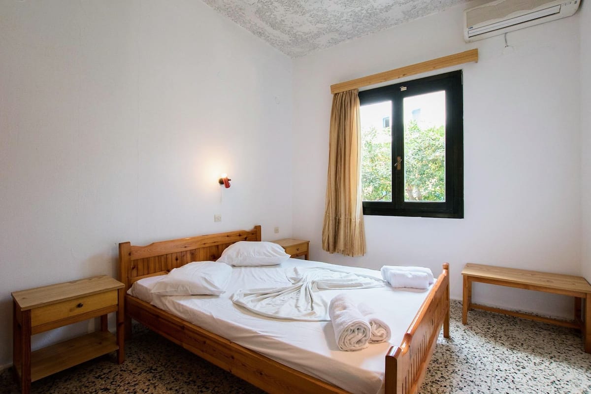 A single-bed room with a wooden headboard, a bedside table, a nightstand, a window with a curtain, and a wall-mounted air conditioner.
