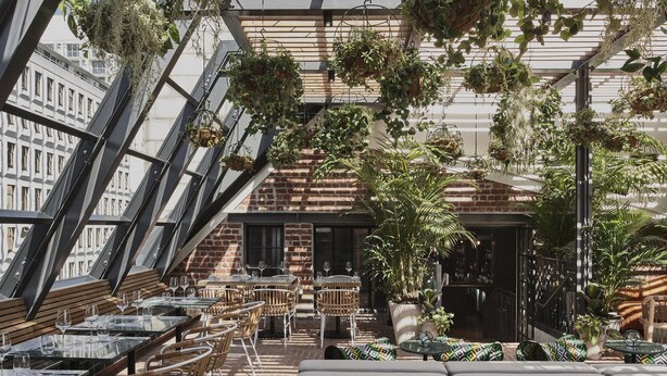 An indoor dining area with high ceilings, exposed brick walls, and hanging plants. Tables and chairs are arranged for dining. - Gorgeous George by Design Hotels (Cape Town)